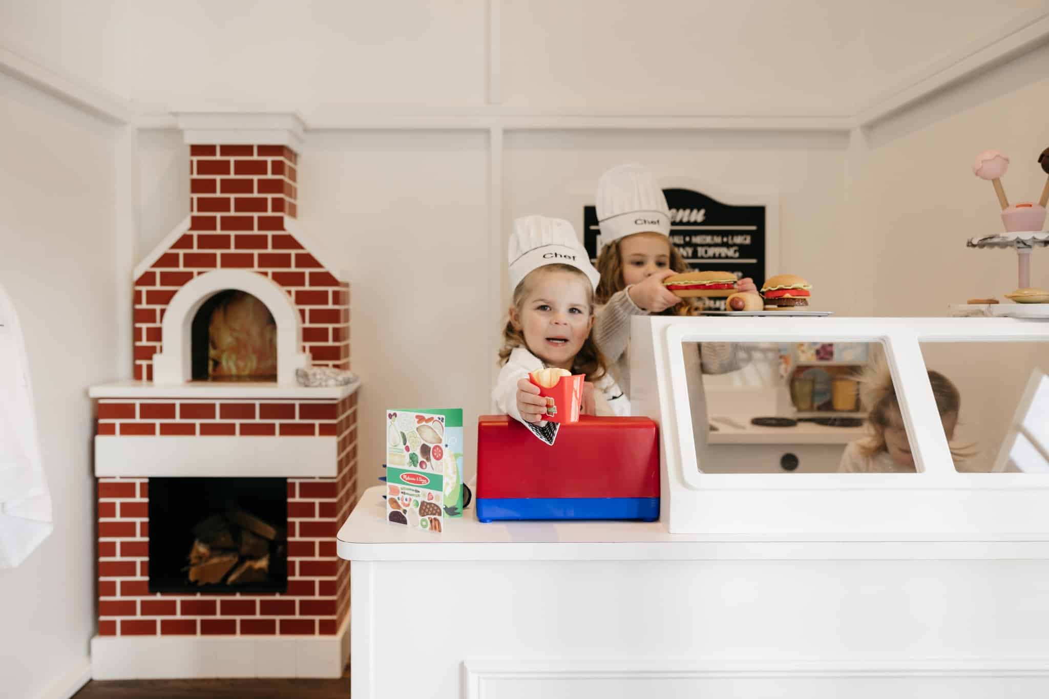 Two children wearing chef hats play in a pretend to cook inside the Main Street Cafe playhouse. In the kitchen there is a brick oven and display counter. A girl is holding toy sandwich. One child is at a play cash register.